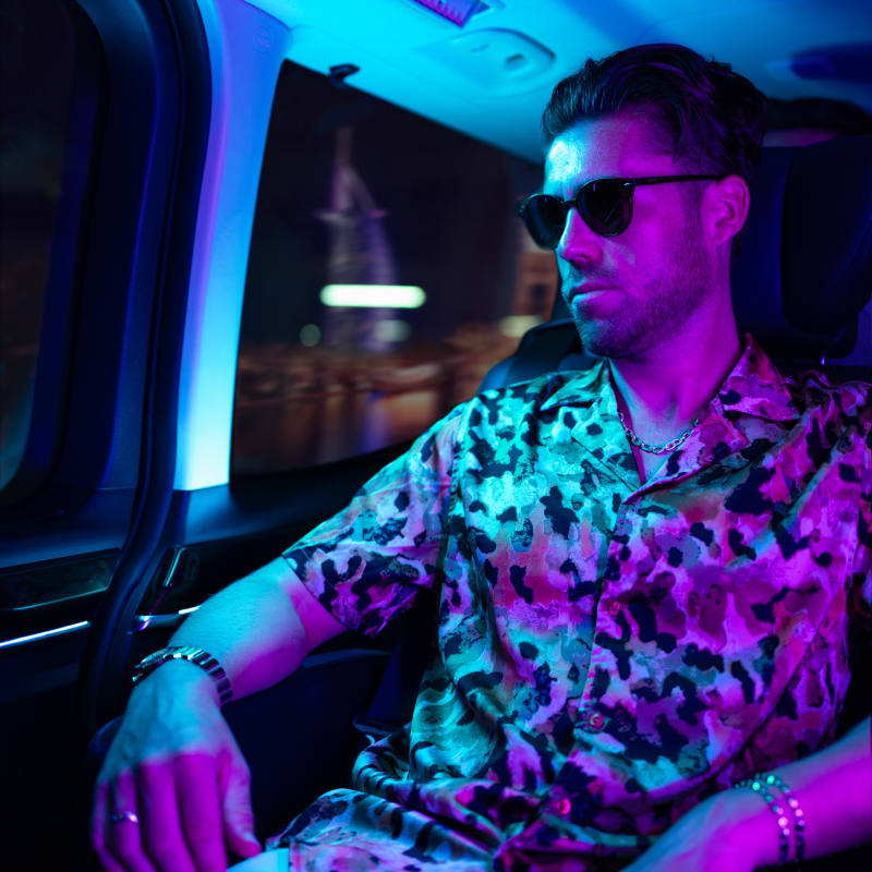 Young man with a floral shirt wearing sunglasses inside a car with purple and blue ambient lighting.